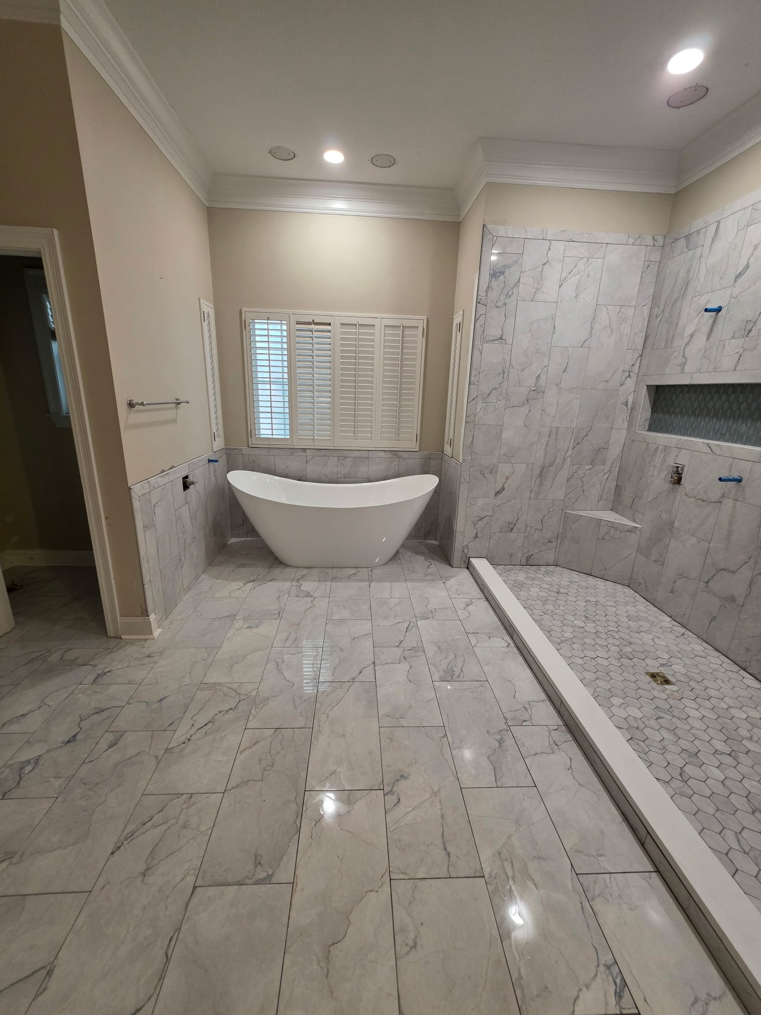 luxury master bathroom with white freestanding soaking tub, marble tile flooring and walls, walk-in shower with glass enclosure, white shutters, recessed lighting, and marble countertops