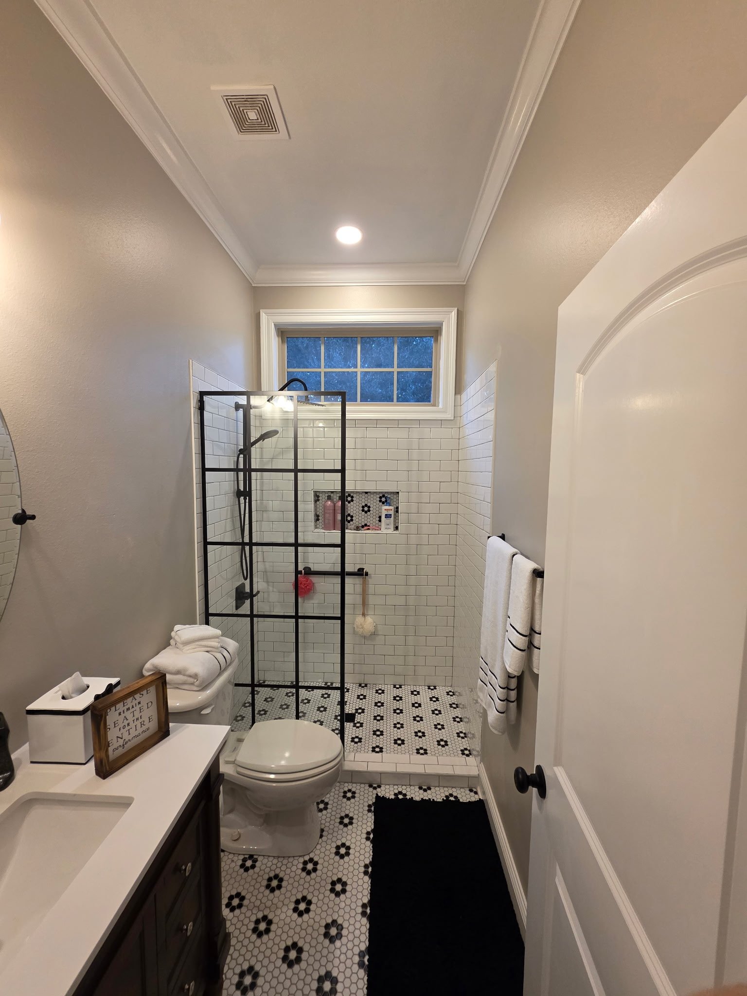 Modern bathroom with white subway tile walls, black framed glass shower enclosure, white vanity, black and white hexagon floor tile, and large window with natural light